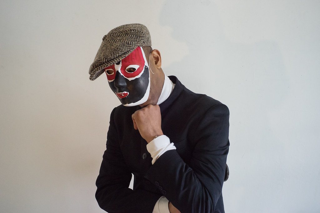 Frantz Lamothe wearing a painted mask and a flat cap, one hand raised to his chin